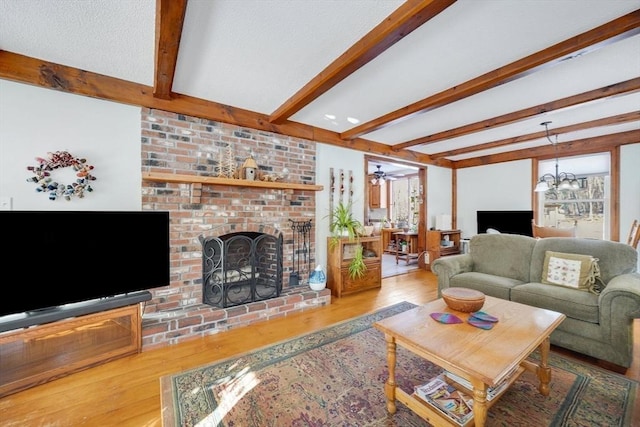 living room featuring ceiling fan with notable chandelier, a brick fireplace, wood-type flooring, and beamed ceiling