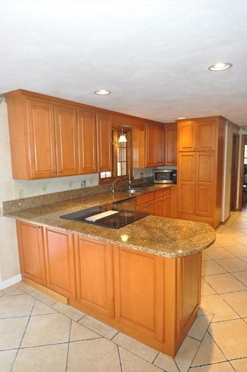 kitchen with stone countertops, brown cabinetry, light tile patterned flooring, stainless steel microwave, and a peninsula