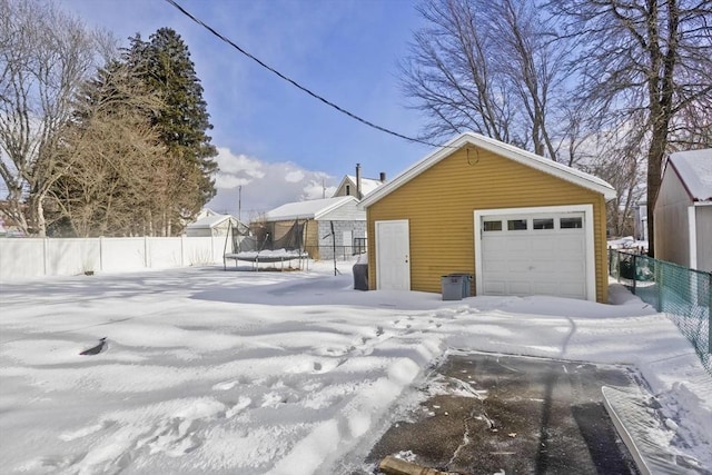 snow covered garage featuring a trampoline, a detached garage, and fence