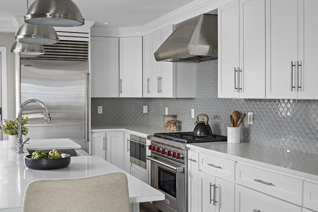 kitchen featuring high end appliances, wall chimney range hood, backsplash, and white cabinetry