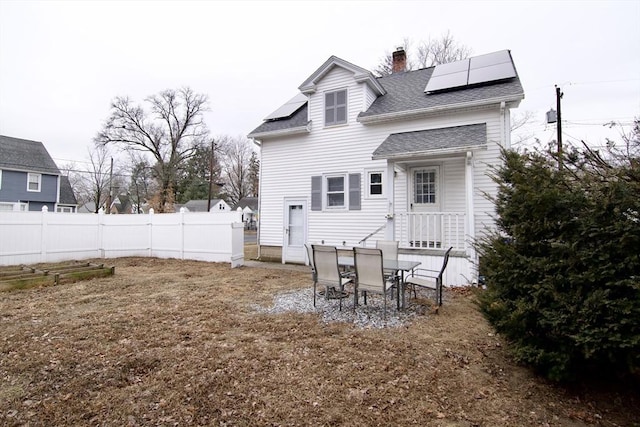 back of house with a chimney, a shingled roof, roof mounted solar panels, fence, and a garden