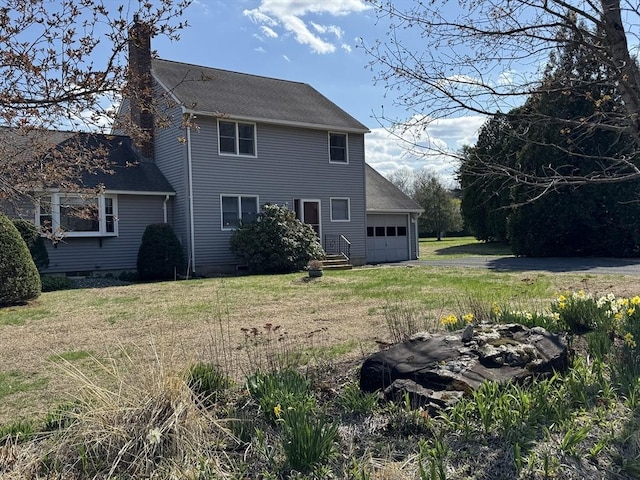 back of house featuring roof with shingles, an attached garage, aphalt driveway, a chimney, and a yard