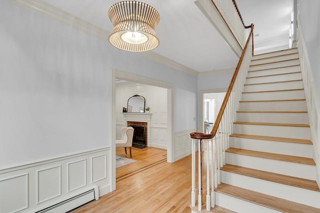 stairway featuring wood finished floors, a wainscoted wall, a baseboard heating unit, crown molding, and a brick fireplace