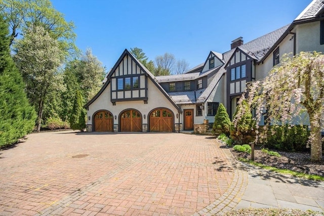 tudor home featuring stucco siding, an attached garage, a chimney, decorative driveway, and stone siding