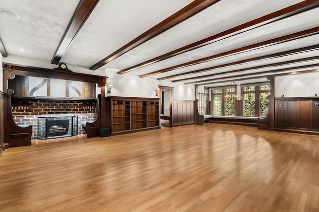 unfurnished living room with beamed ceiling, a brick fireplace, wood finished floors, and a wainscoted wall