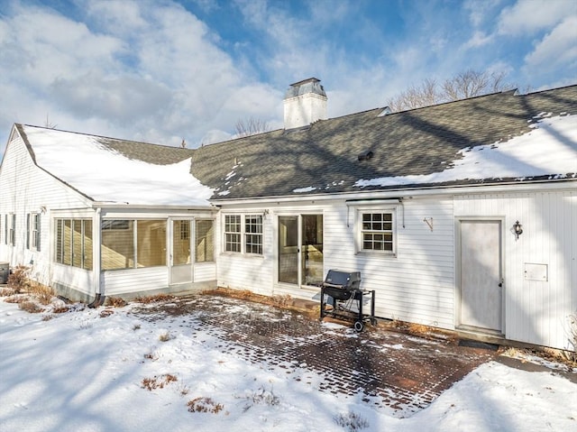 snow covered house featuring a sunroom, roof with shingles, and a chimney