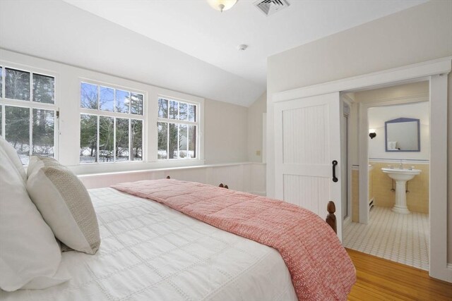 bedroom featuring ensuite bath, a wainscoted wall, wood finished floors, vaulted ceiling, and visible vents