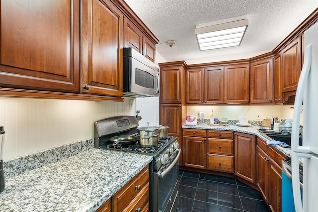 kitchen with stainless steel appliances, dark tile patterned flooring, a textured ceiling, light stone counters, and brown cabinetry