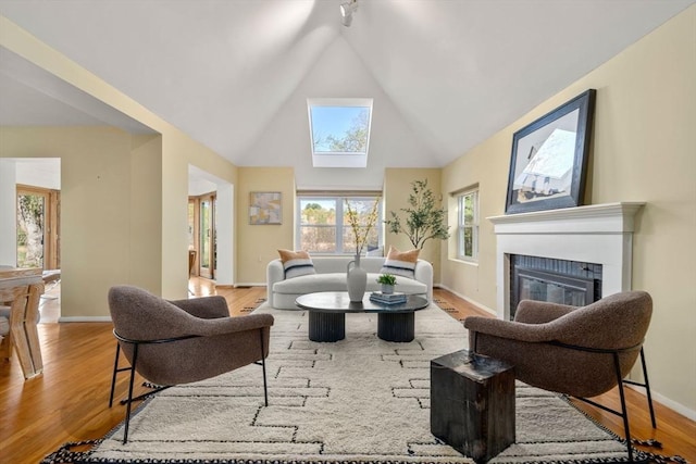 living area featuring a skylight, wood finished floors, high vaulted ceiling, baseboards, and a glass covered fireplace