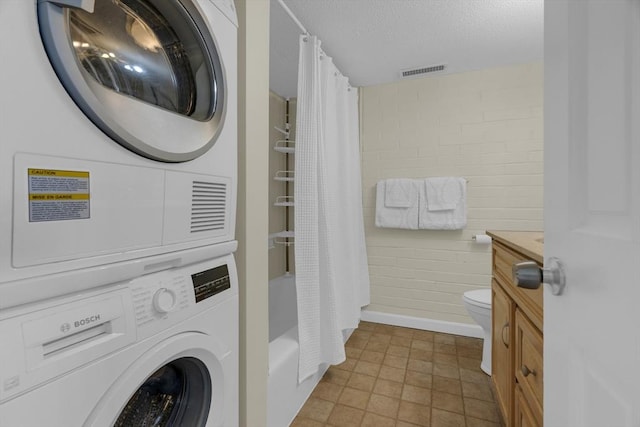 laundry area with stacked washer / drying machine, brick wall, and a textured ceiling