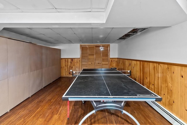 playroom featuring dark wood-style flooring, wooden walls, a paneled ceiling, a baseboard heating unit, and a wainscoted wall