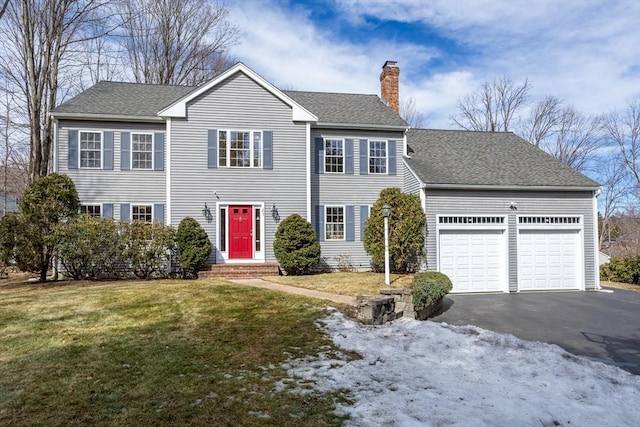 colonial house featuring a shingled roof, a front lawn, aphalt driveway, a chimney, and a garage