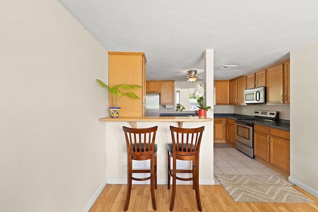 kitchen featuring stainless steel appliances, a kitchen breakfast bar, a ceiling fan, brown cabinetry, and light wood-type flooring