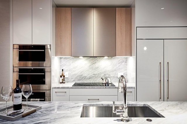 kitchen with backsplash, black electric stovetop, sink, light stone counters, and stainless steel double oven