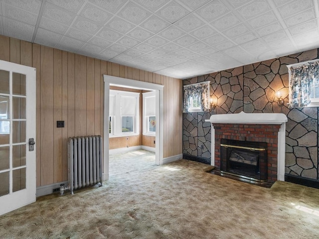 unfurnished living room featuring radiator, carpet, a fireplace, wood walls, and baseboards