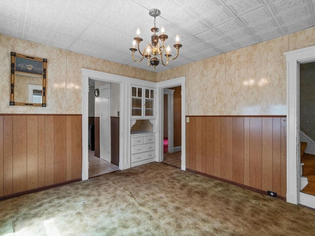 unfurnished dining area featuring wainscoting, a chandelier, carpet flooring, wood walls, and an ornate ceiling
