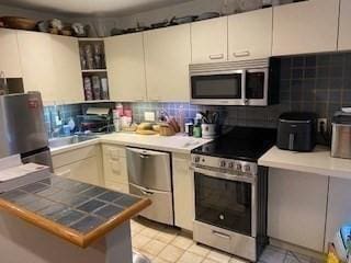 kitchen featuring open shelves, decorative backsplash, appliances with stainless steel finishes, and tile countertops