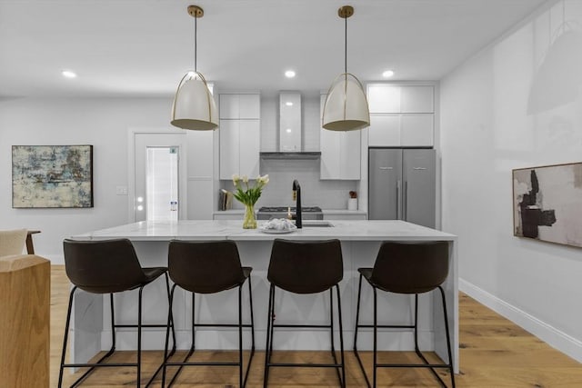 kitchen featuring white cabinets, appliances with stainless steel finishes, wall chimney range hood, hanging light fixtures, and a center island with sink