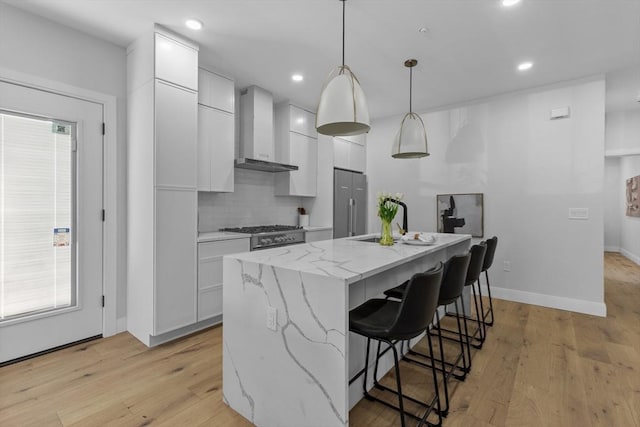 kitchen featuring a center island with sink, hanging light fixtures, light stone countertops, white cabinets, and wall chimney exhaust hood