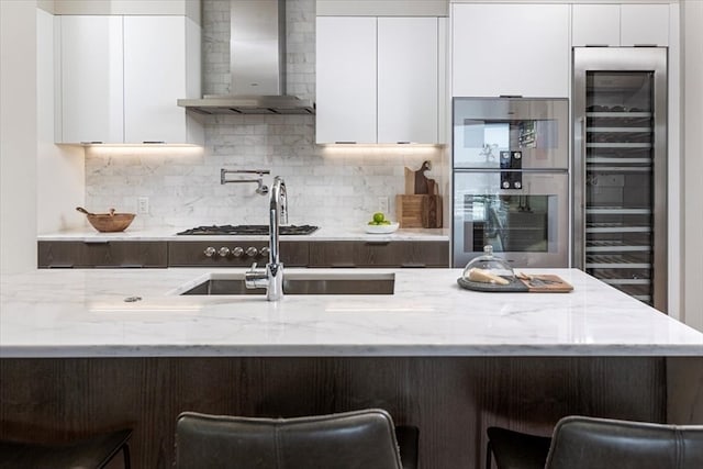 kitchen featuring white cabinetry, double oven, light stone countertops, wall chimney exhaust hood, and tasteful backsplash