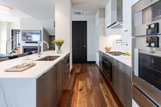 kitchen featuring appliances with stainless steel finishes, sink, dark wood-type flooring, wall chimney exhaust hood, and dark brown cabinetry