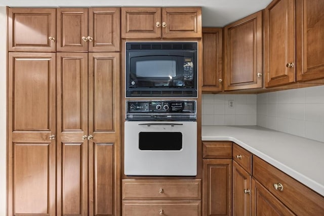 kitchen featuring white oven, brown cabinetry, black microwave, tasteful backsplash, and light countertops