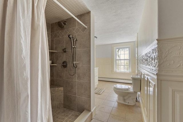 full bath with tile patterned flooring, wainscoting, toilet, a baseboard heating unit, and a tile shower