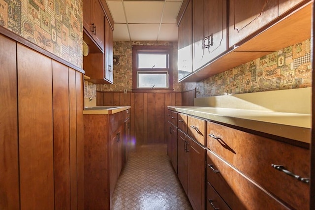 kitchen featuring brown cabinetry, a paneled ceiling, wood walls, and a sink