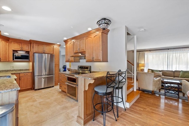 kitchen featuring a breakfast bar, brown cabinets, appliances with stainless steel finishes, open floor plan, and under cabinet range hood