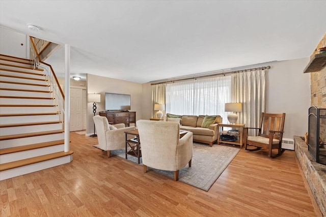 living area featuring stairs, a baseboard heating unit, a stone fireplace, and light wood-style flooring