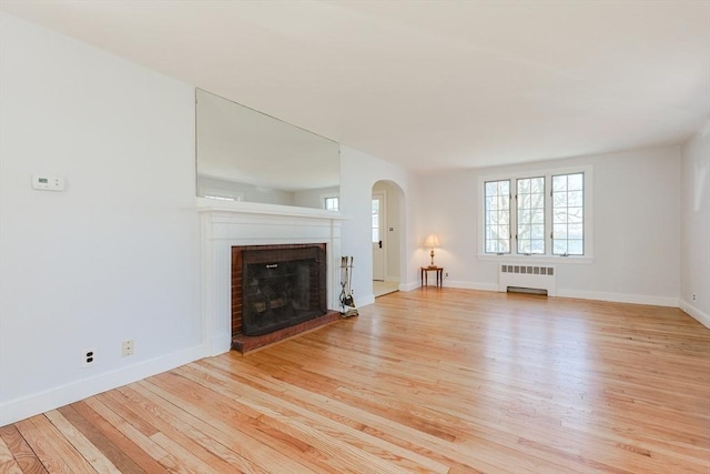 unfurnished living room featuring radiator, a fireplace, baseboards, arched walkways, and light wood finished floors