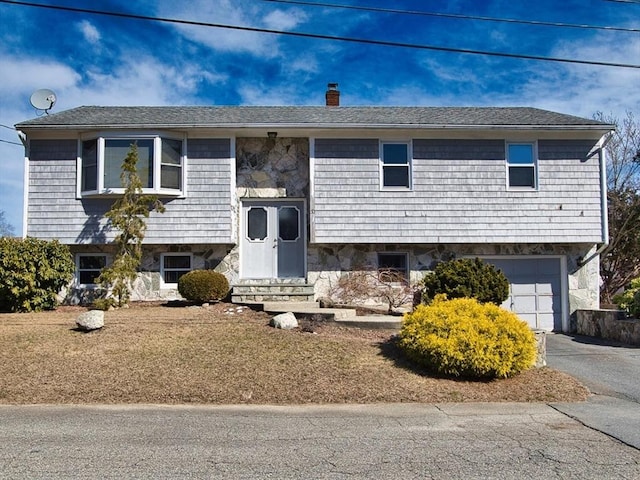 bi-level home featuring an attached garage, entry steps, a chimney, stone siding, and driveway