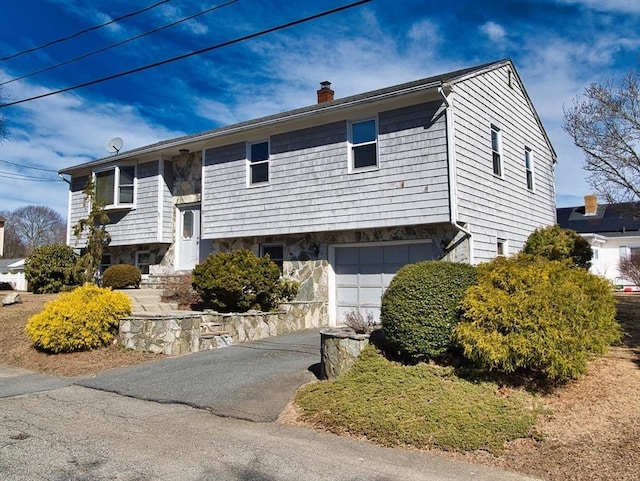 bi-level home featuring aphalt driveway, stone siding, a chimney, and a garage