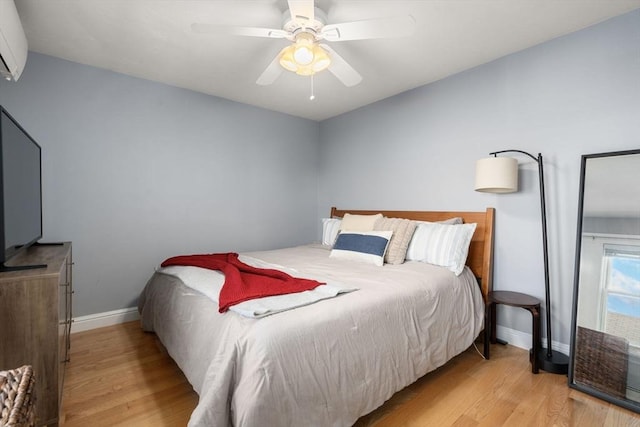 bedroom with ceiling fan, a wall mounted AC, light wood-type flooring, and baseboards