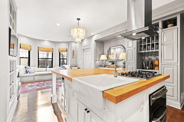 kitchen with wooden counters, island exhaust hood, hanging light fixtures, a kitchen island with sink, and stainless steel gas stovetop