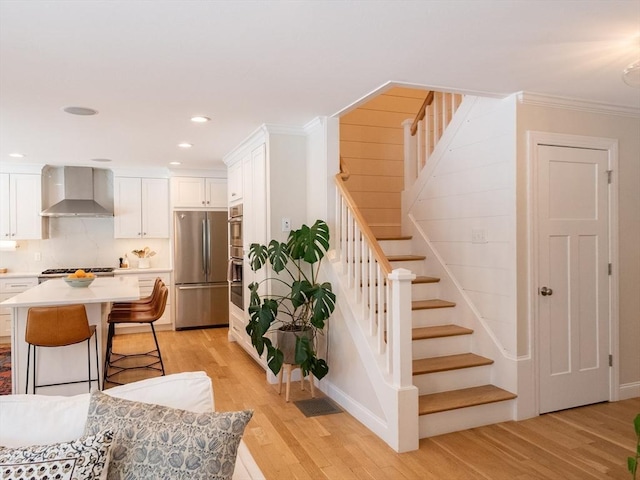 kitchen featuring white cabinets, wall chimney range hood, stainless steel appliances, a kitchen breakfast bar, and ornamental molding