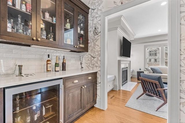 bar featuring dark brown cabinets, tasteful backsplash, light hardwood / wood-style floors, ornamental molding, and beverage cooler