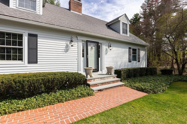 doorway to property featuring a lawn, a chimney, and a shingled roof