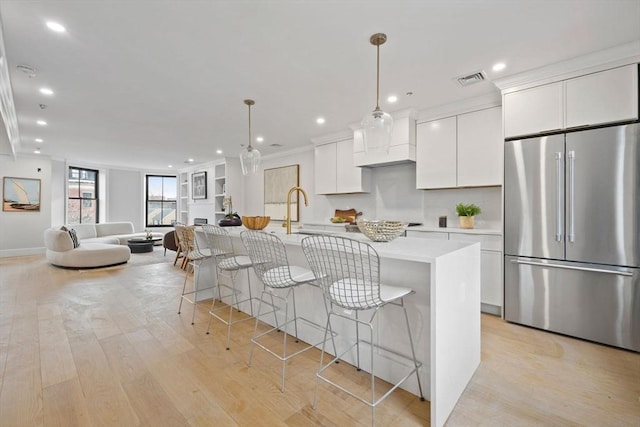 kitchen featuring white cabinets, decorative light fixtures, stainless steel fridge, light wood-type flooring, and a center island with sink