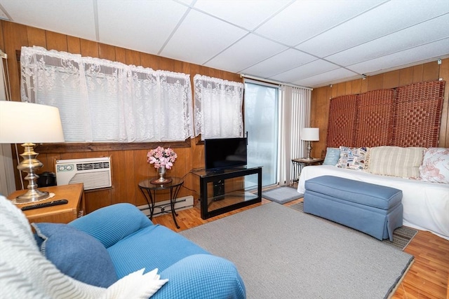 living room featuring hardwood / wood-style floors, a baseboard heating unit, wooden walls, a wall mounted AC, and a drop ceiling