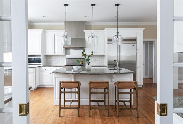 kitchen featuring wall chimney exhaust hood, a center island with sink, crown molding, and built in appliances