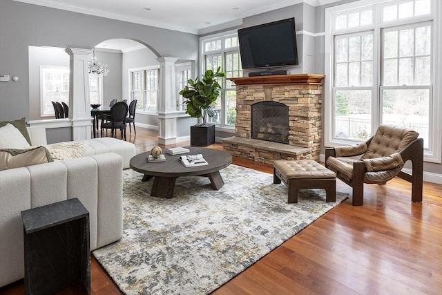 living room with arched walkways, a stone fireplace, wood finished floors, ornamental molding, and ornate columns