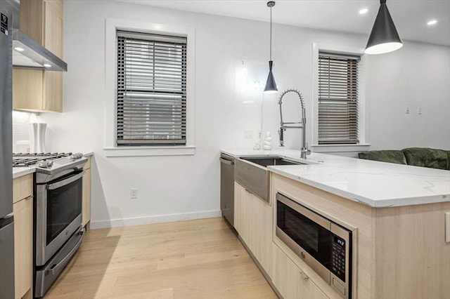 kitchen with light brown cabinets, wall chimney exhaust hood, hanging light fixtures, appliances with stainless steel finishes, and light wood-type flooring