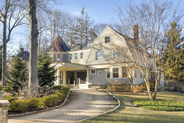 shingle-style home featuring a porch, decorative driveway, a gambrel roof, and a chimney