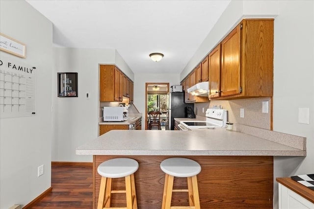 kitchen featuring a breakfast bar, under cabinet range hood, white appliances, a peninsula, and brown cabinetry