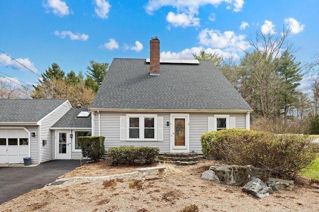 cape cod-style house with roof mounted solar panels, a chimney, aphalt driveway, an attached garage, and roof with shingles