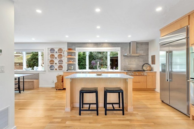 kitchen with wall chimney range hood, appliances with stainless steel finishes, light brown cabinetry, and light wood finished floors