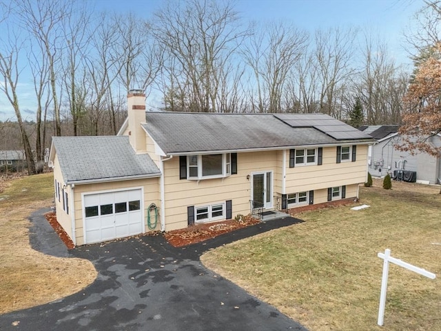 bi-level home featuring solar panels, a front lawn, aphalt driveway, a chimney, and an attached garage