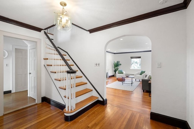 staircase with crown molding, arched walkways, wood finished floors, baseboards, and a chandelier