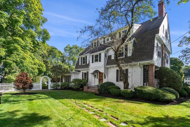 view of front facade with brick siding, a shingled roof, a gambrel roof, a front yard, and fence